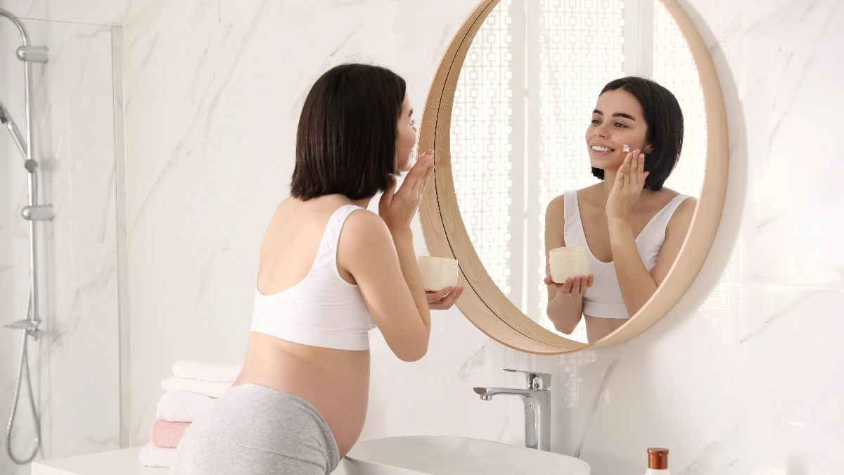  Schwangere Frau schaut in den Spiegel und trägt Gesichtspflege auf 